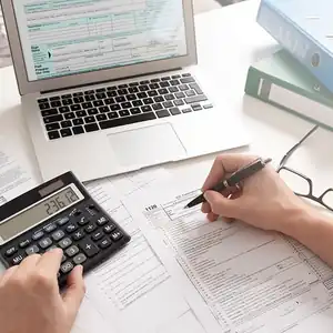 A person is filling out a tax form with a pen in their right hand and a calculator in their left hand, while their laptop is open in front of them displaying additional tax forms.