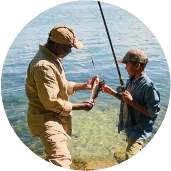 A father and son with brown skin stand in front of a lake as the son shows off a fish he caught.