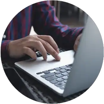 The hands of a man with tan skin and a checkered button-down shirt work on a laptop.