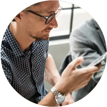 A young white man with glasses sits and looks at his phone.