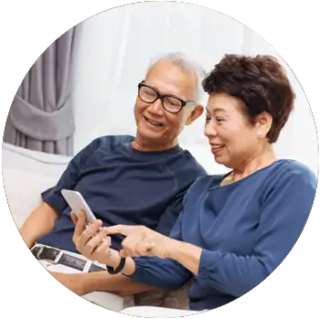 A senior-aged Asian couple smile while they sit on a couch and look at a mobile device.
